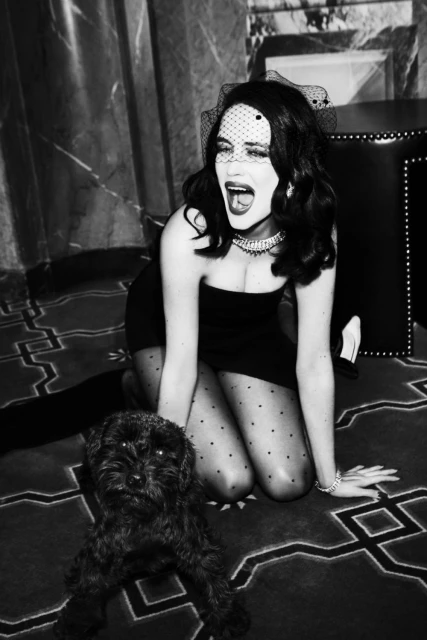 Woman in black dress, veil, and tights kneels on a patterned carpet with a black dog. Dramatic lighting and black and white photography.