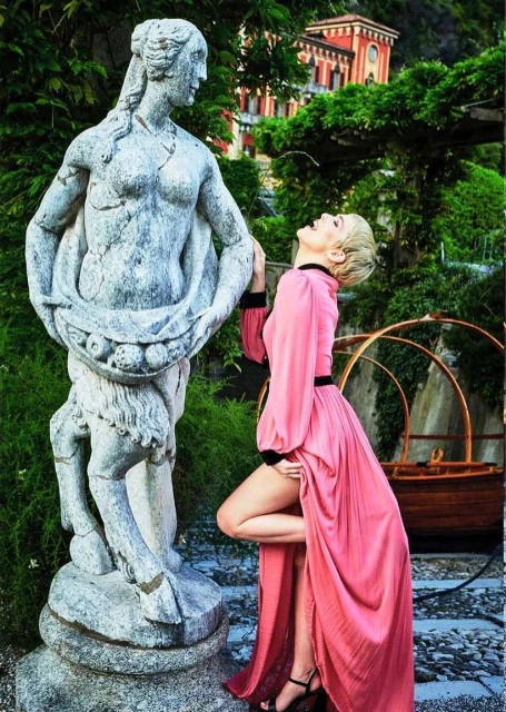 Woman in flowing pink dress poses next to a weathered stone statue outdoors with a building and trees in the blurred background.