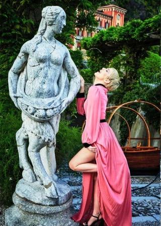 Woman in flowing pink dress poses next to a weathered stone statue outdoors with a building and trees in the blurred background.