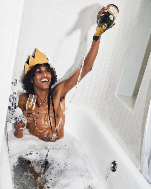 Person in bathtub pouring champagne, wearing a crown, jewelry, and smiling, with white tiled walls in a bright indoor setting.