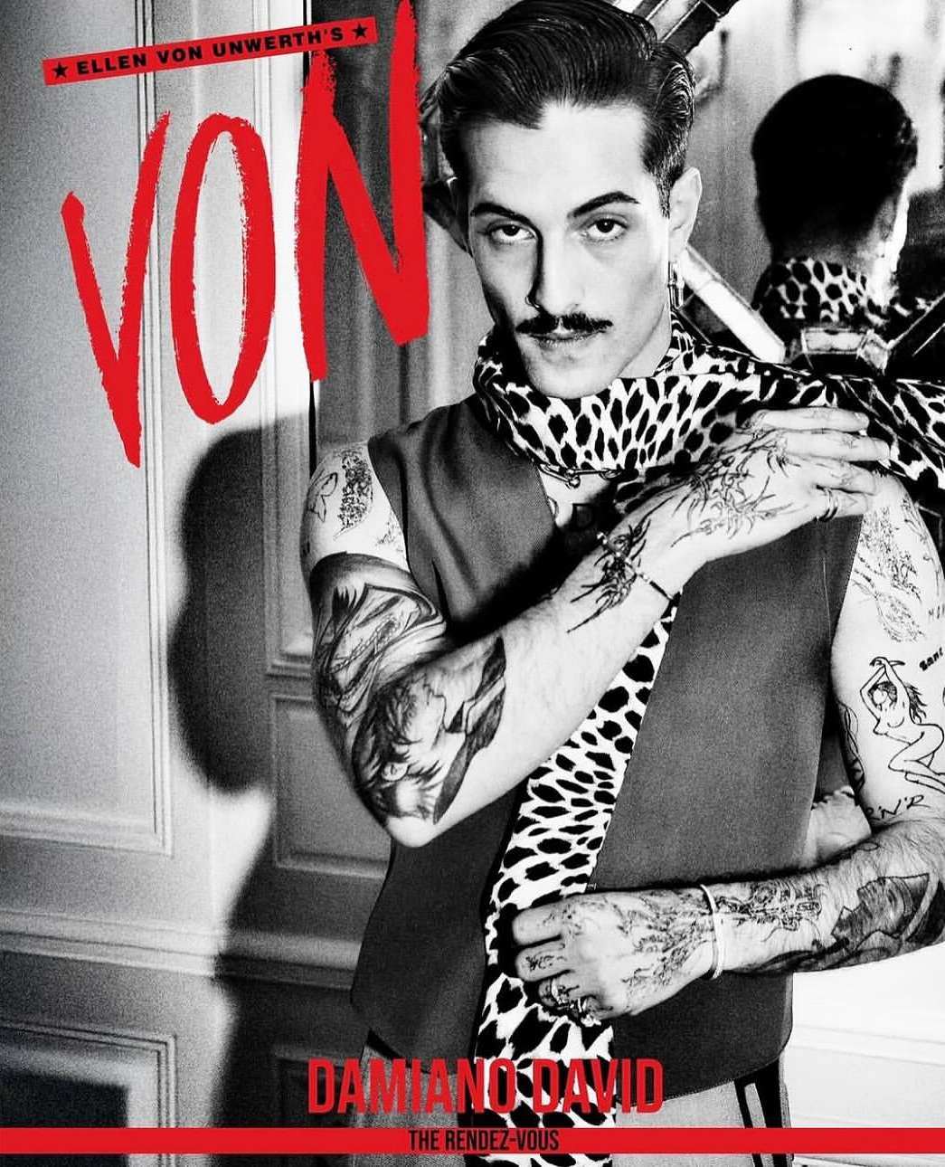 Fashion portrait of Damiano David with a mustache and tattoos, wearing a vest and animal print scarf, posing against a paneled wall.