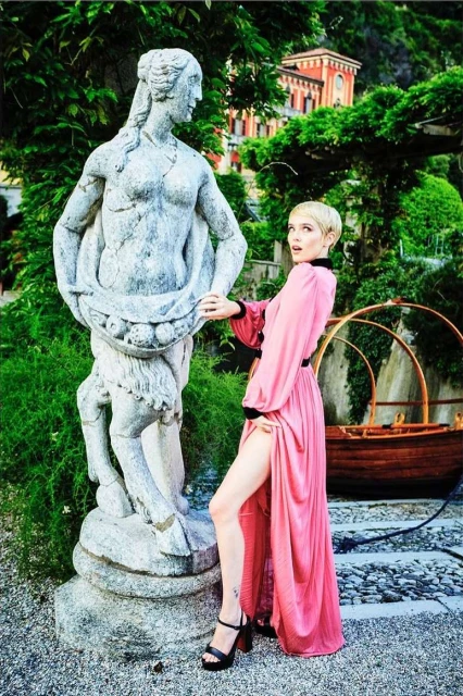 Adult female in a pink dress poses next to a human-animal form marble statue in a lush green outdoor setting with a red brick building.