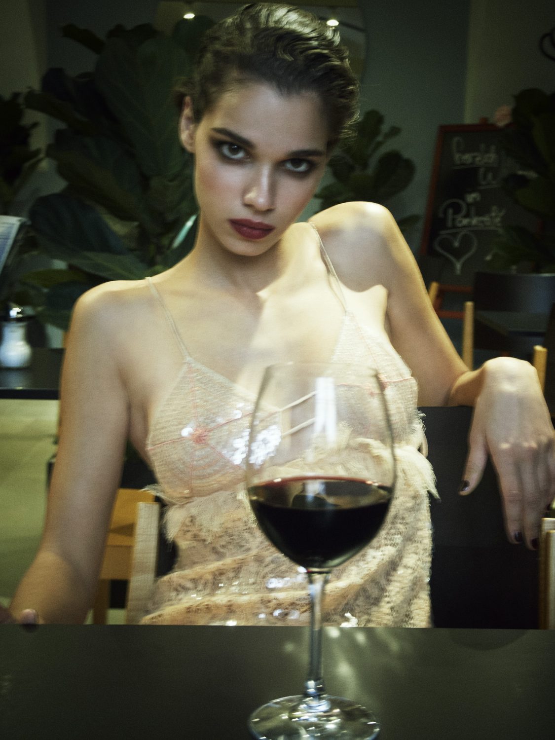 Indoor portrait of a model seated at a table, featuring a wine glass with red wine and a backdrop of leafy green plants.