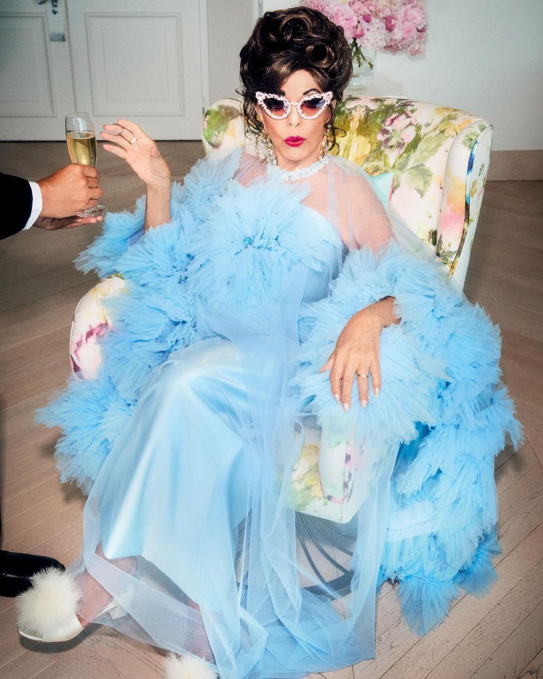 Elegant older woman in a light blue dress and feathered robe sits in a floral chair, holding a champagne glass, offered by a server.