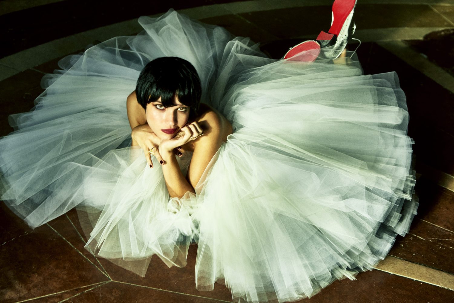 A woman with dark hair poses while lying on a tiled floor in a white tulle dress, paired with red-soled heels in a fashion shot.