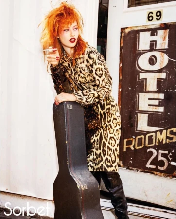 Woman with red hair in leopard coat holding glass, guitar case, standing near Hotel sign, fashion photography.
