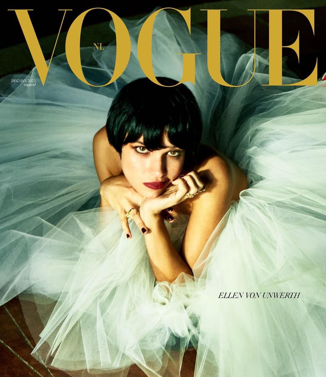 Fashion magazine cover featuring a woman with dark hair and red lipstick wearing a white tulle dress, posing with gold rings.