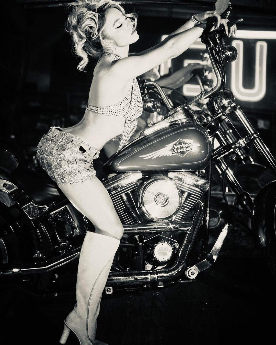 Blonde adult female poses on a Harley Davidson motorcycle in a black-and-white photo with an urban backdrop and shiny chrome details.