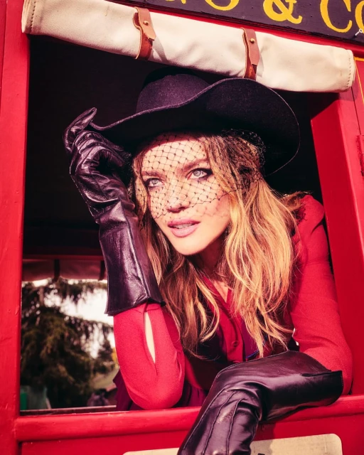 Woman in red dress, black hat and gloves poses inside a red cabin looking out of the window with trees visible in the distance.