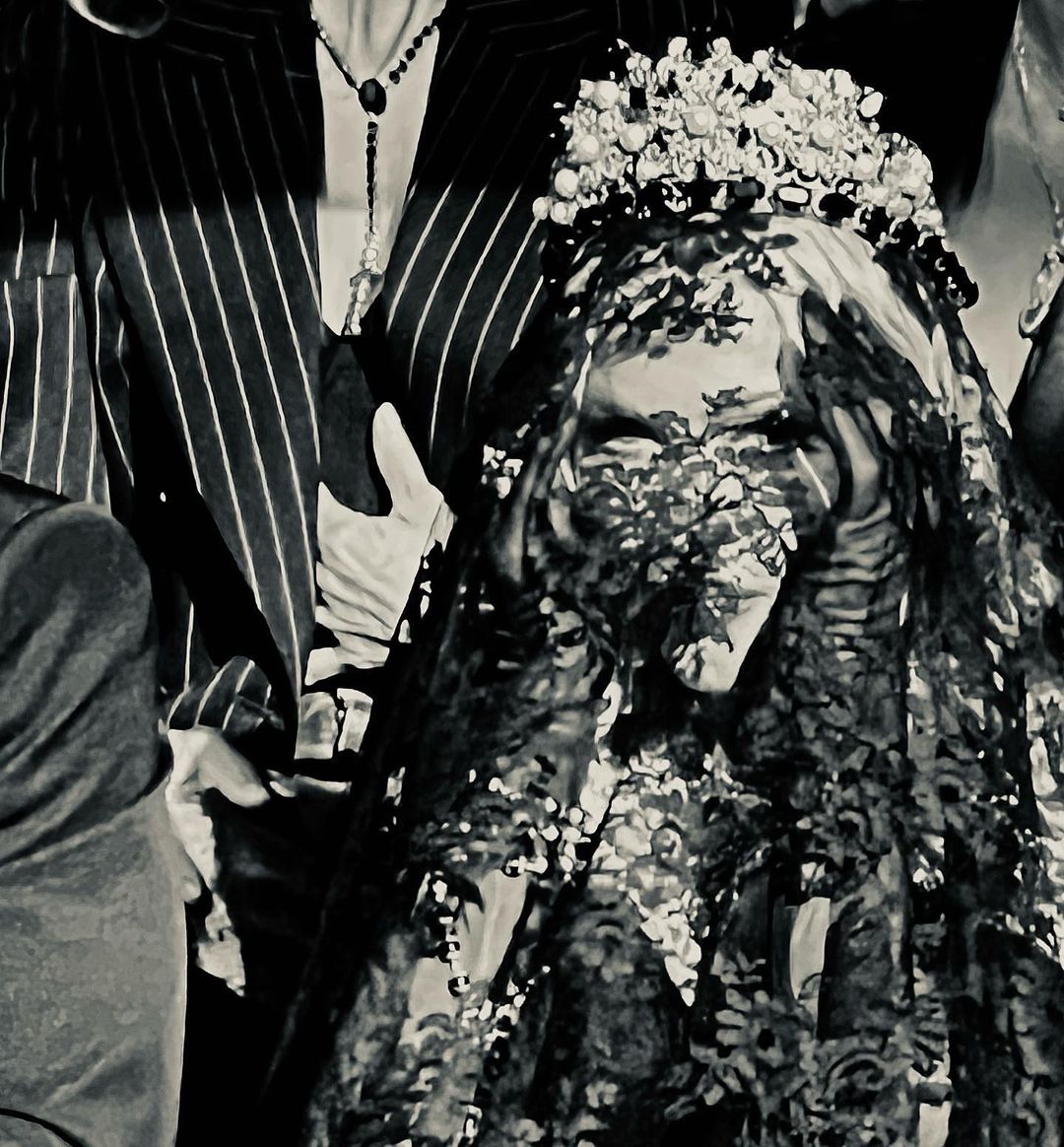 A bride wearing a lace veil and flower crown stands surrounded by people in a black and white image, captured in a close-up.