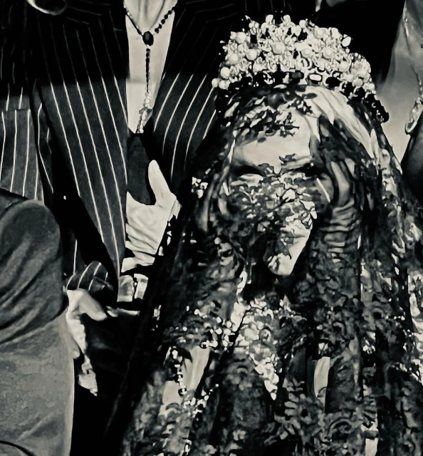 A bride wearing a lace veil and flower crown stands surrounded by people in a black and white image, captured in a close-up.
