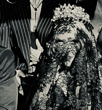 A bride wearing a lace veil and flower crown stands surrounded by people in a black and white image, captured in a close-up.