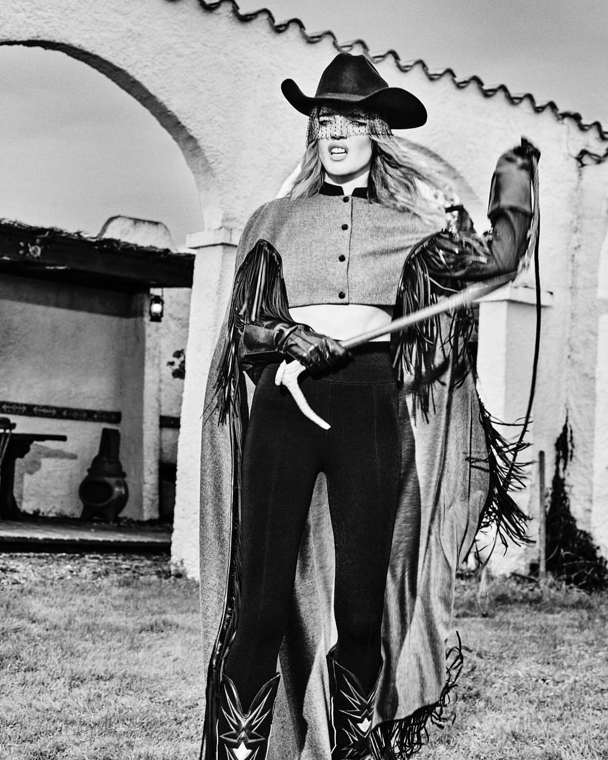 Black-and-white portrait of a woman in a cowboy hat and outfit holding a whip, in an outdoor grassy environment with stucco archway.
