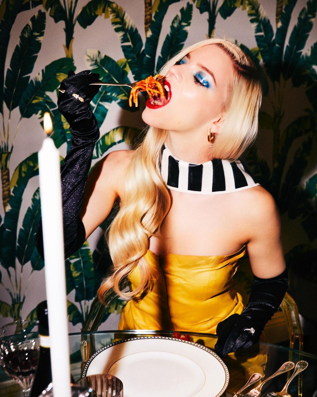 Blonde woman in yellow dress eating spaghetti with dramatic eye makeup and palm leaf wallpaper backdrop, elegant dining scene.