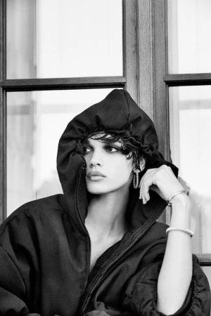 Monochrome portrait of a woman wearing a dark hooded jacket and hoop earrings against a window.