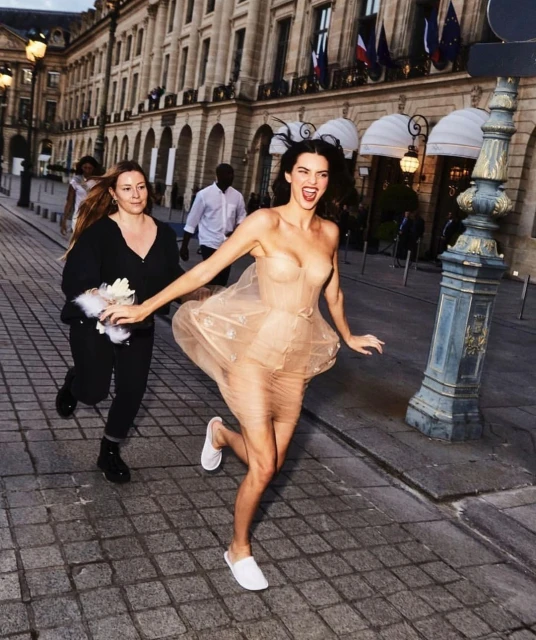 Kendall Jenner runs with a friend in a tulle dress and white slippers in an urban environment with building architecture.