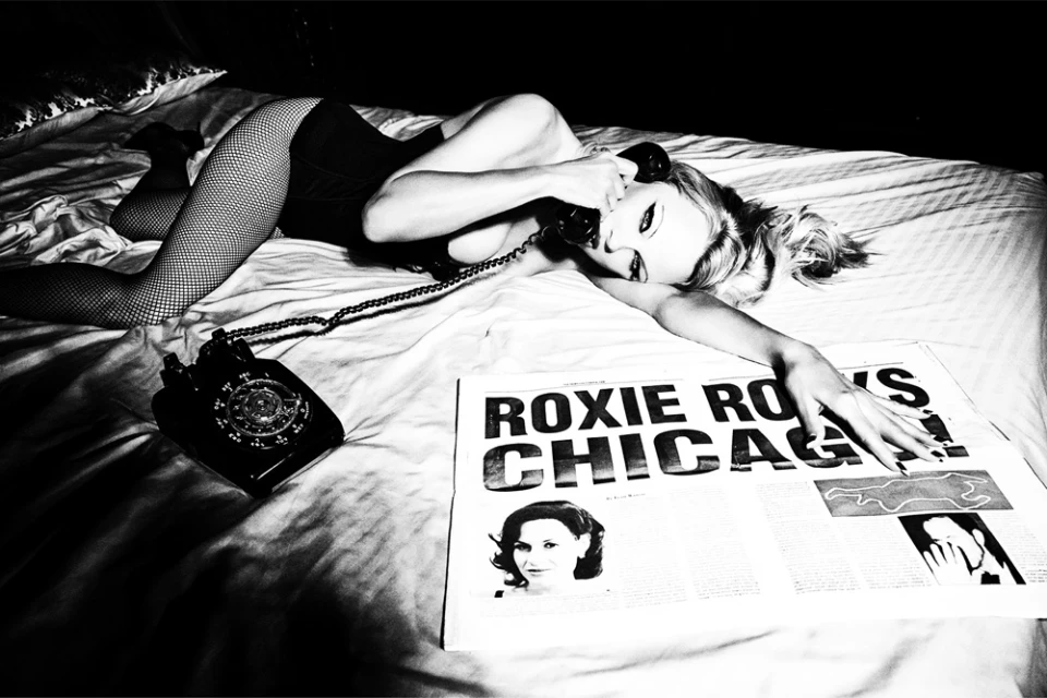 Monochrome portrait of a woman in fishnets on a bed, holding a rotary phone, beside a newspaper, bathed in dramatic lighting.