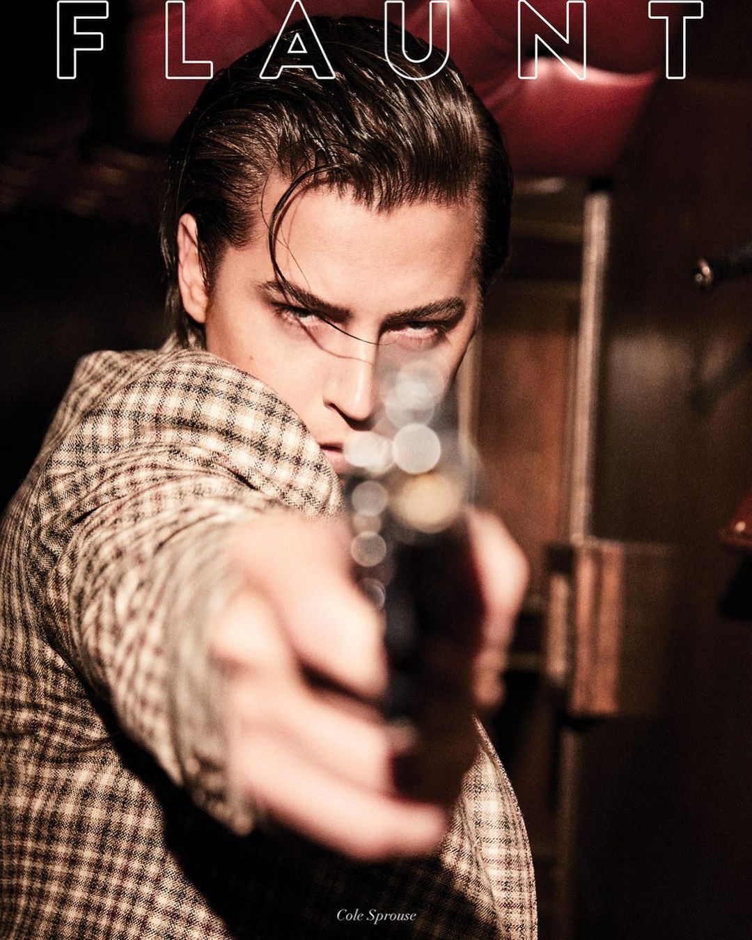 Portrait of a man in a patterned jacket pointing a handgun directly at the camera lens, with "FLAUNT" magazine cover text.