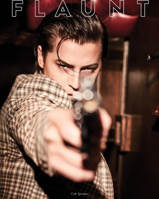 Portrait of a man in a patterned jacket pointing a handgun directly at the camera lens, with "FLAUNT" magazine cover text.