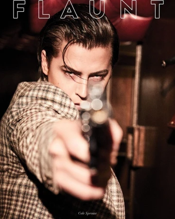 Portrait of a man in a patterned jacket pointing a handgun directly at the camera lens, with "FLAUNT" magazine cover text.
