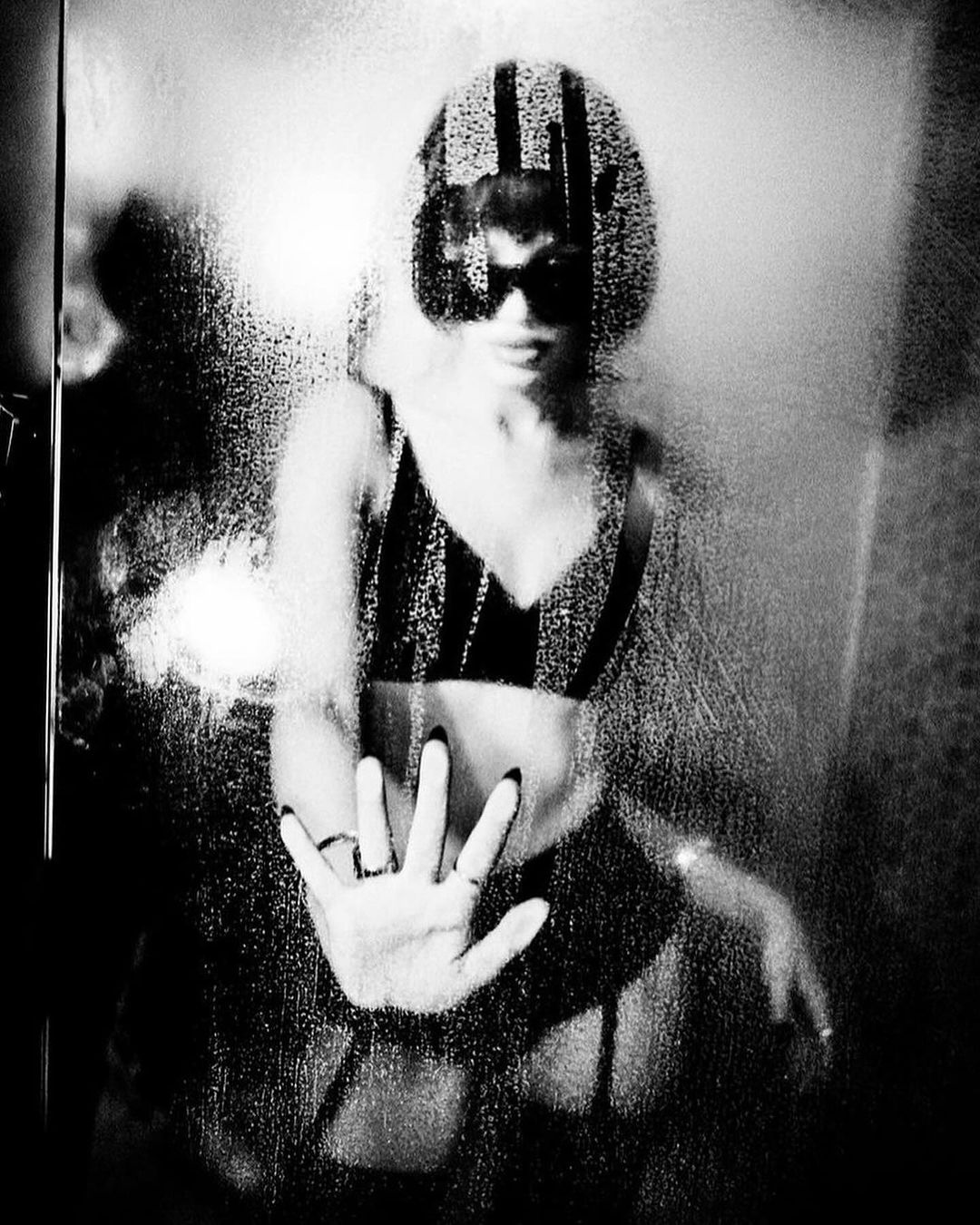 Monochrome image of an adult female in lingerie behind a wet glass surface, obscuring her face and body, creating a stylized effect.