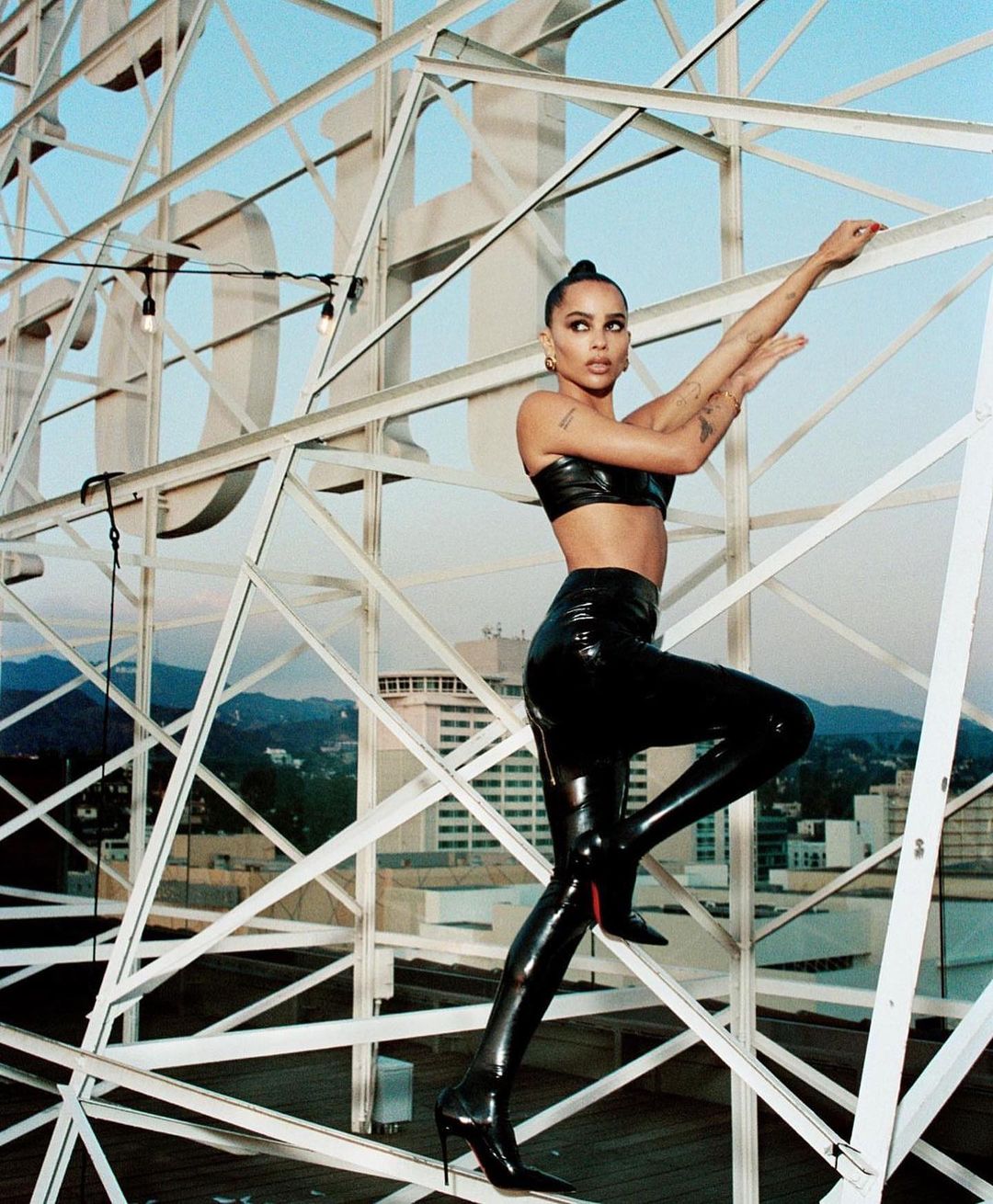Woman in black vinyl outfit poses on Hollywood sign structure against a blue sky backdrop. The shot features buildings in the background.