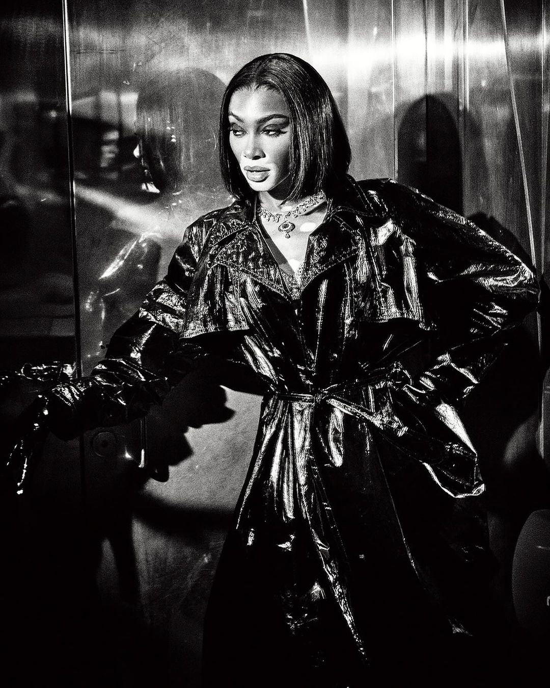 Black and white portrait of a woman wearing a shiny patent leather trench coat and necklace, shot by Ellen Von Unwerth.