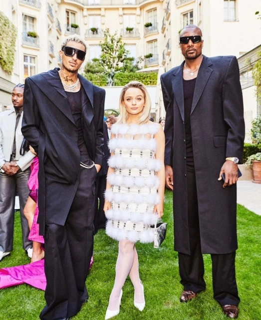 Two men and a woman pose outdoors in designer clothing. The men wear black coats and sunglasses, and the woman wears a white feathered dress.