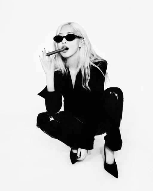 Blonde woman in black suit and sunglasses squats indoors, holding a cigar. Black and white portrait on a white seamless backdrop.