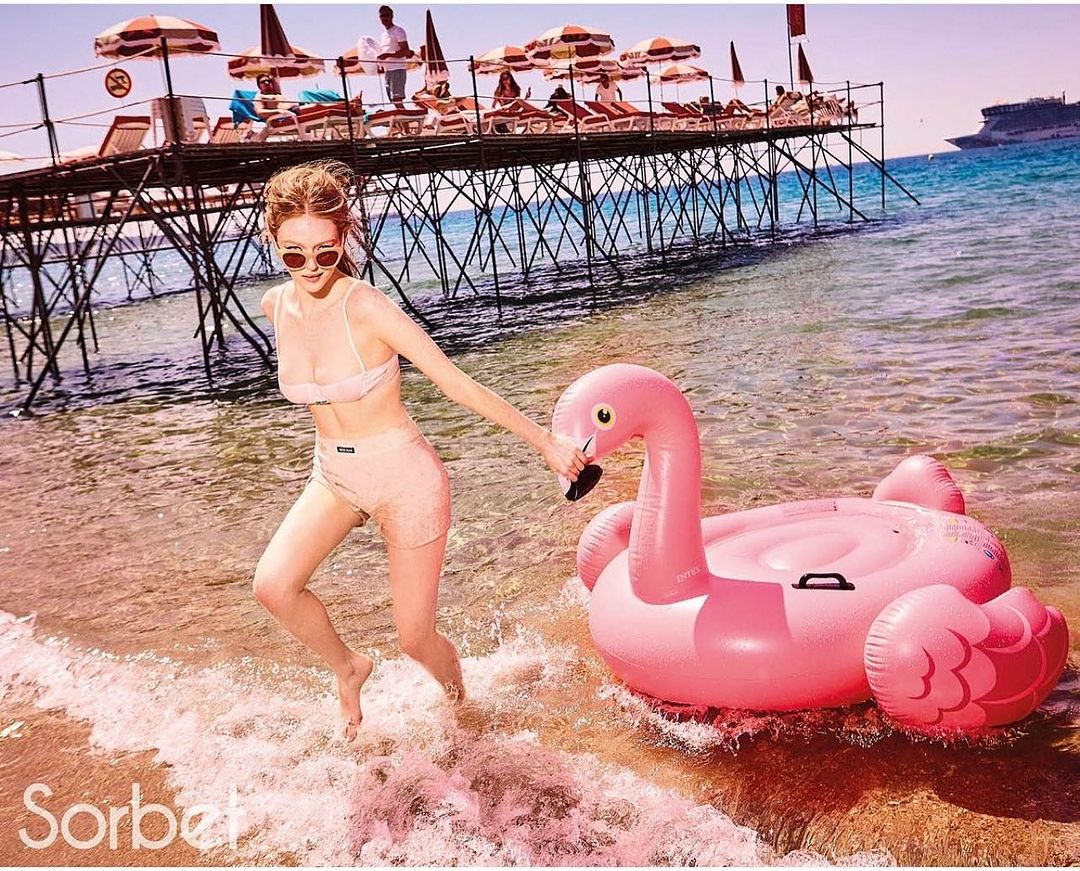 A woman in a bikini runs at the beach with a pink flamingo inflatable toy near a pier and the ocean during a sunny day.