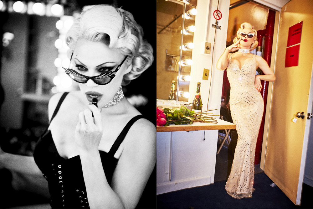 Woman in glamorous attire applying lipstick in a backstage dressing room with a table, roses, and champagne under bright vanity lights.