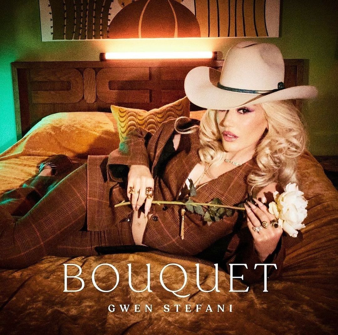 Gwen Stefani poses on a bed wearing a cowboy hat and brown plaid suit, holding a bouquet of white flowers in a portrait studio shot.