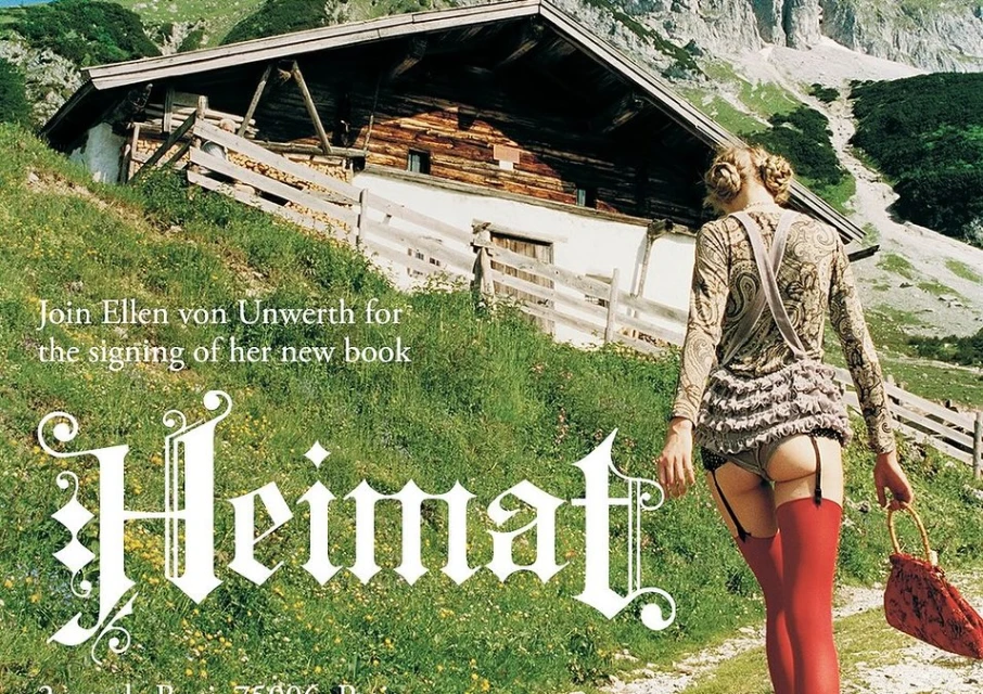 Adult female in lingerie, long-sleeved shirt, and suspenders walks along a grass path with a house and mountain backdrop.