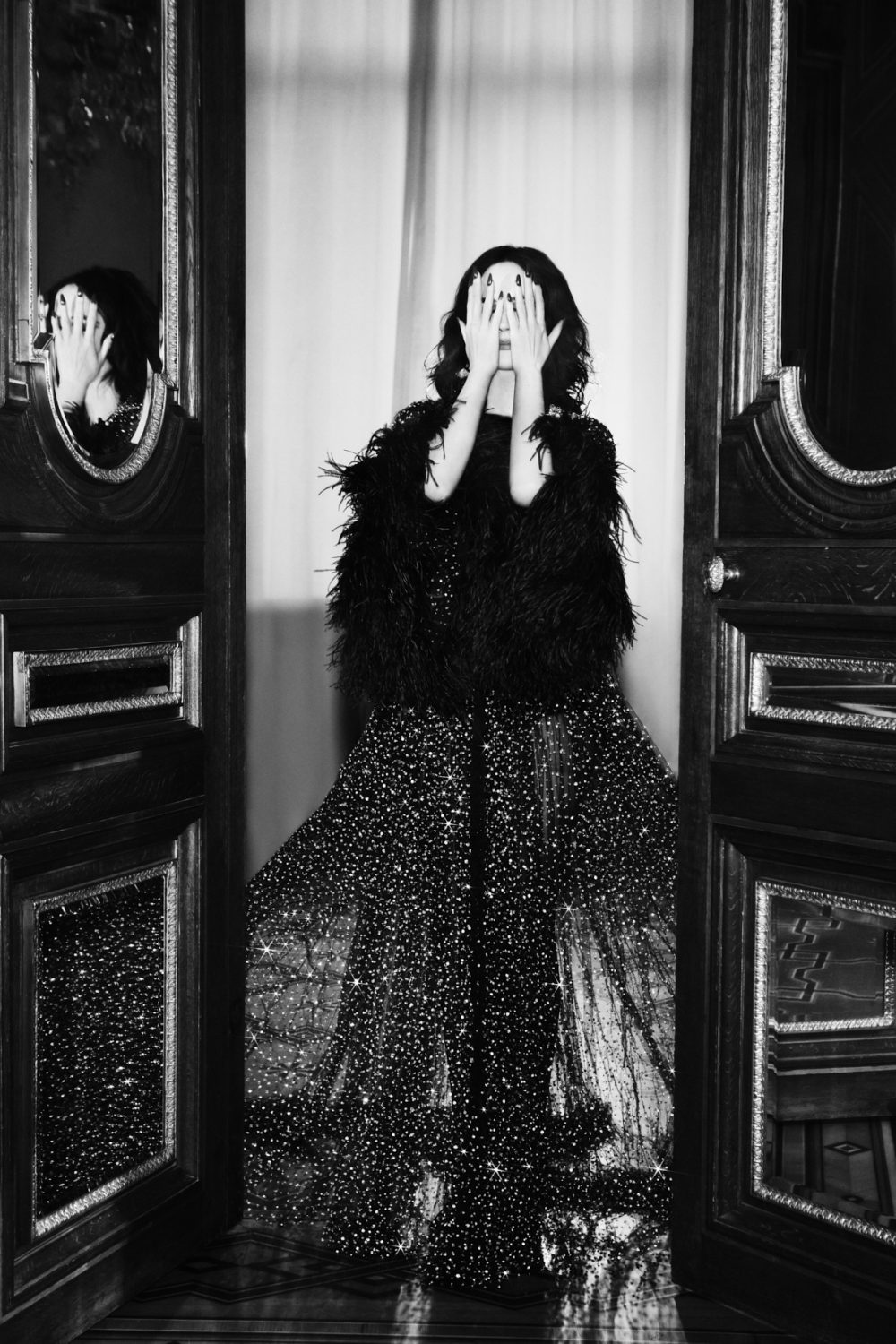 Monochrome portrait of a woman in an ornate dress, her face obscured by her hands, standing in a doorway with reflective mirrors.