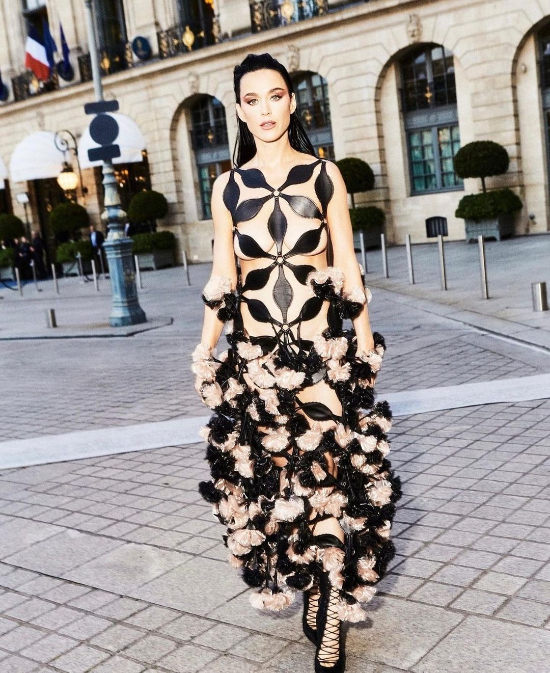 Woman in elaborate black-and-beige dress walks on stone-paved urban street with building in the background, French flag is visible.