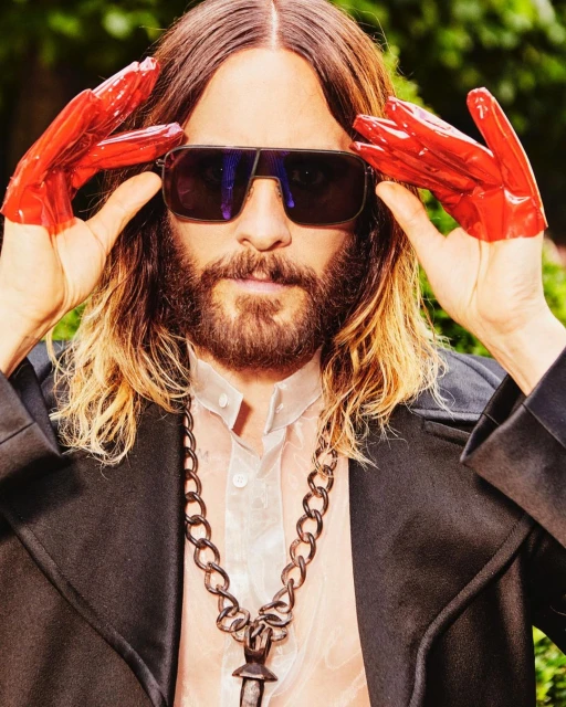 Close-up portrait of a man with long hair and a beard wearing sunglasses and red gloves, set against a blurred green background.