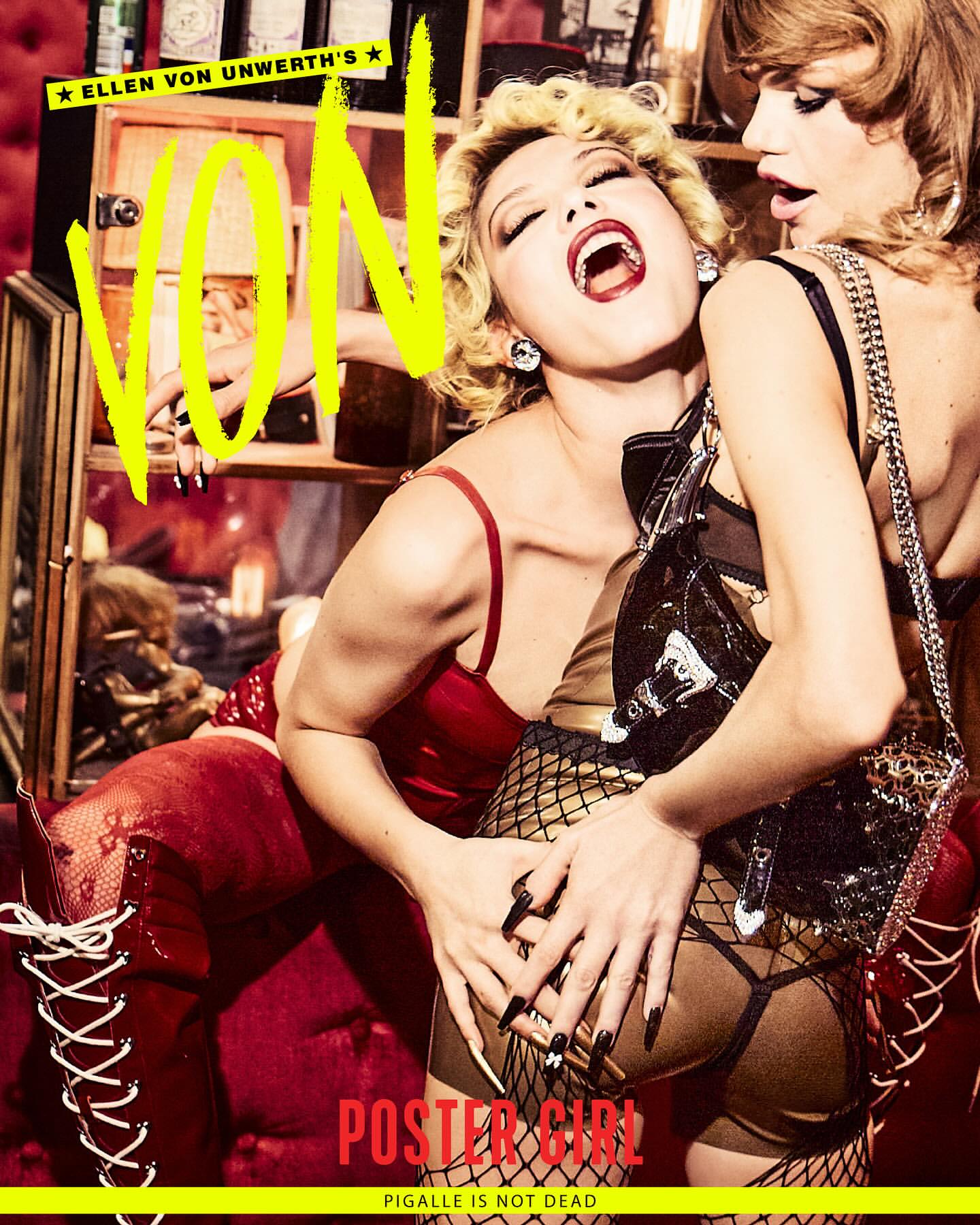 Two women pose together indoors for a magazine cover; one touches the other’s rear end as both pose in dark lighting.