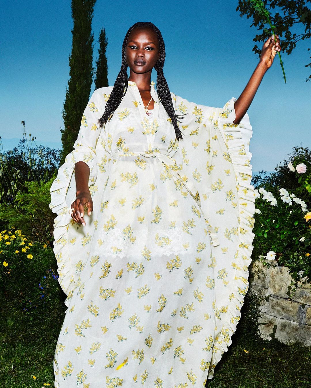 Elegant Black woman in a flowing, floral-patterned white dress in a garden setting, holding a leafy branch, fashion editorial.