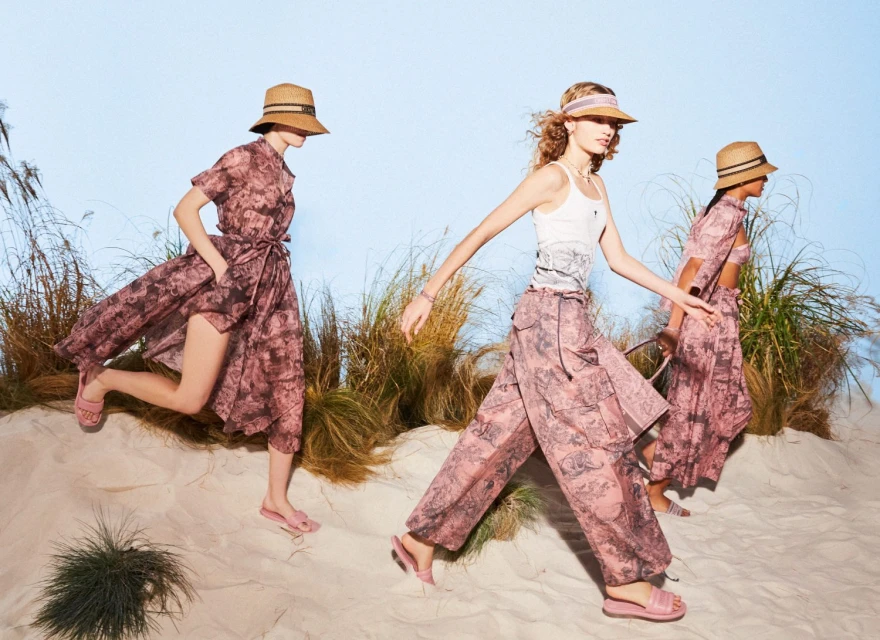 Three women in patterned dresses and hats walk on a sandy surface against a blue sky. Fashion shot with outdoor setting.