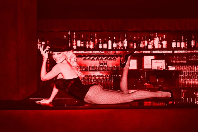 Woman in corset and hat posing on bar counter under red monochrome lighting with liquor bottles in background