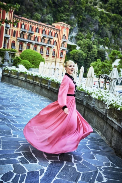 Woman in a pink gown twirls along a stone path with white flowers, a historic building, and a hillside in the background.