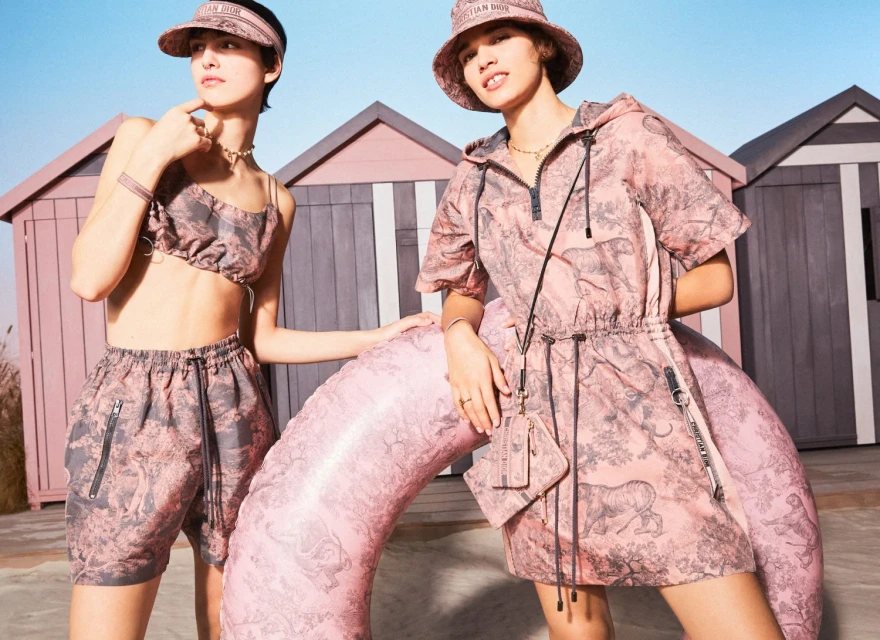 Two women in matching pink and gray print outfits pose with a pink inflatable tube in front of striped beach huts, branded with Christian Dior.