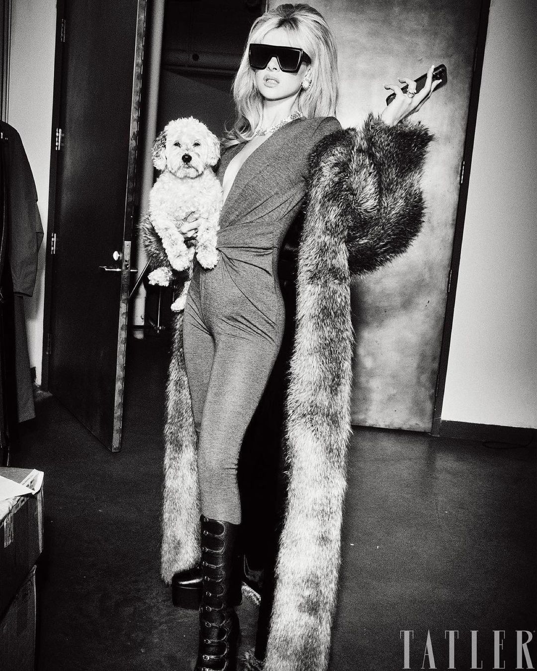 Blonde woman in a bodysuit and knee-high boots holds a small dog in an indoor photo studio setting with a faux fur stole.