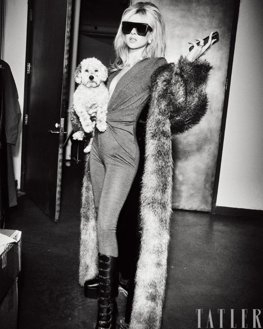Blonde woman in a bodysuit and knee-high boots holds a small dog in an indoor photo studio setting with a faux fur stole.