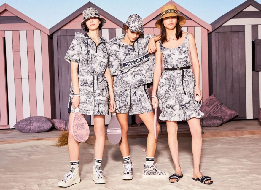 Three fashion models pose on a sandy beach wearing patterned outfits with beach huts in the background, for a Dior campaign.