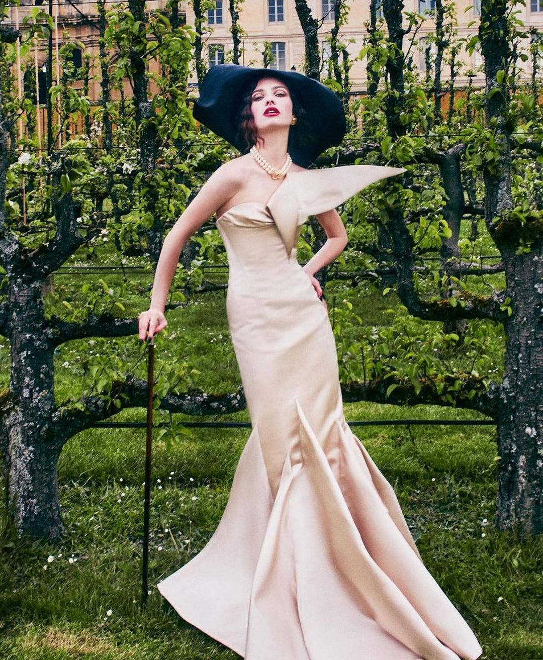 Elegant woman in strapless gown, wide-brimmed hat and cane, standing in a verdant garden setting near blossoming trees and old building.