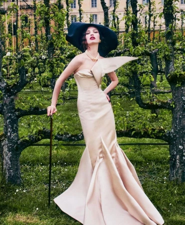 Elegant woman in strapless gown, wide-brimmed hat and cane, standing in a verdant garden setting near blossoming trees and old building.
