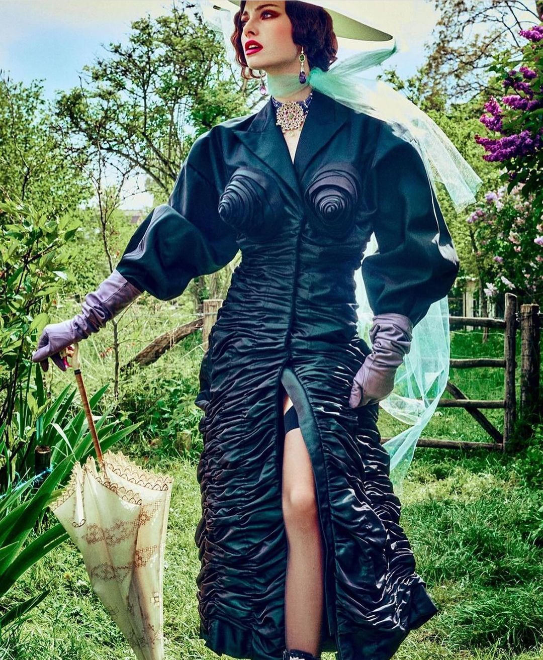 Elegant woman in couture dark dress and wide-brim hat with veil poses in a green garden with an umbrella, creating a fashion editorial photograph.