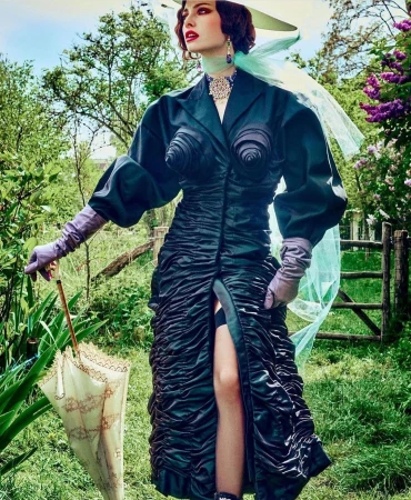 Elegant woman in couture dark dress and wide-brim hat with veil poses in a green garden with an umbrella, creating a fashion editorial photograph.