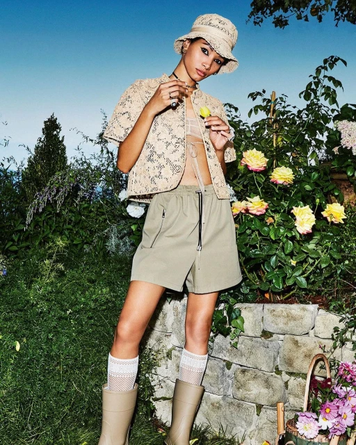 A tan-skinned model in a beige lace top, olive shorts, and bucket hat poses in a garden with roses, wearing boots and patterned socks.